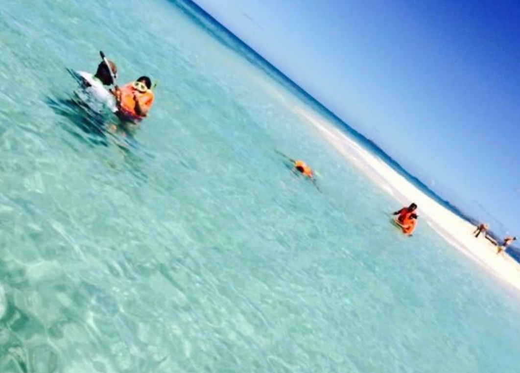Guests in orange life vests snorkelling in waist-deep clear water beside a white sand bar
