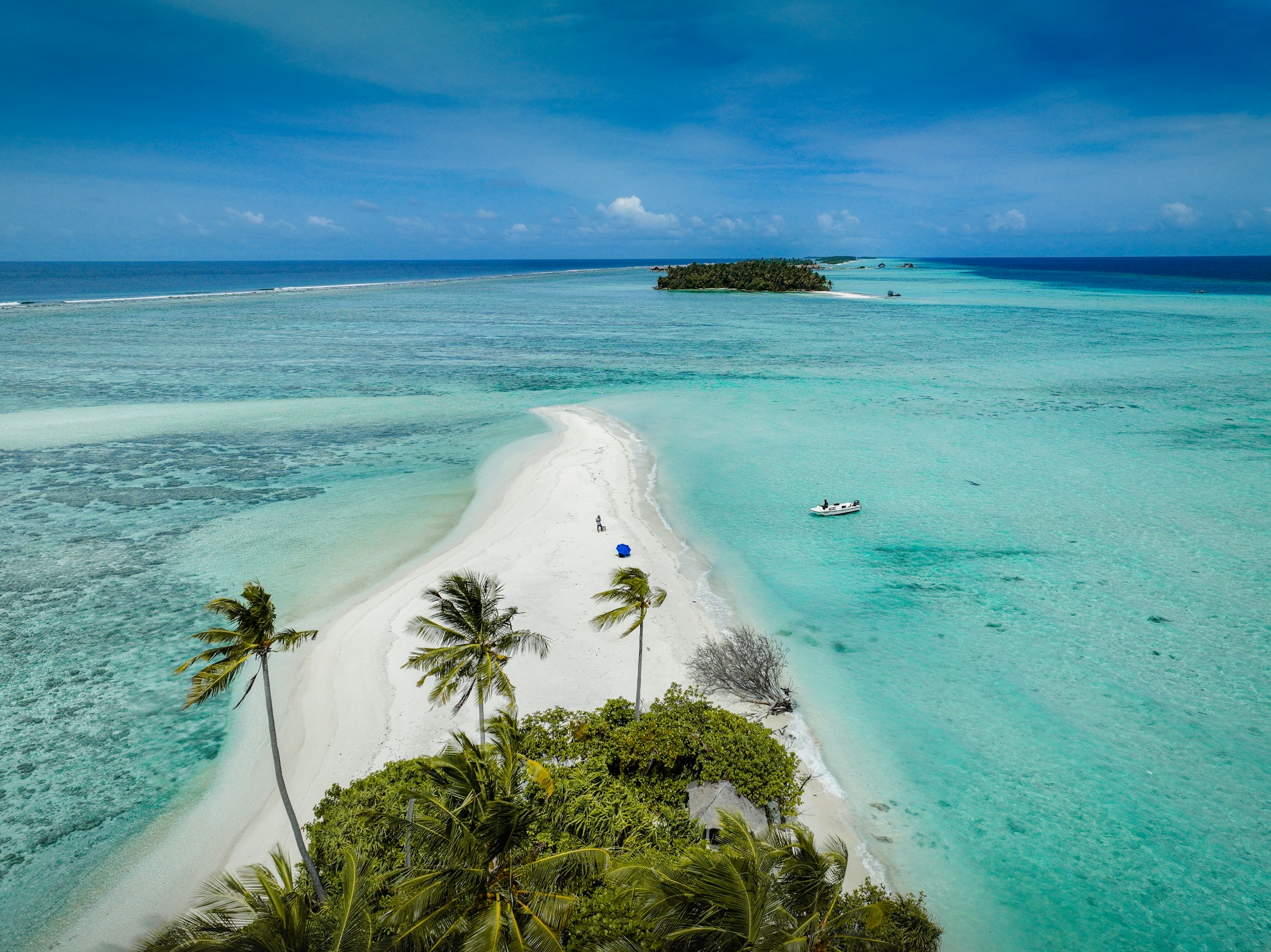 Aerial view of a white sandbar with a few palms, a small boat anchored in the turquoise shallows